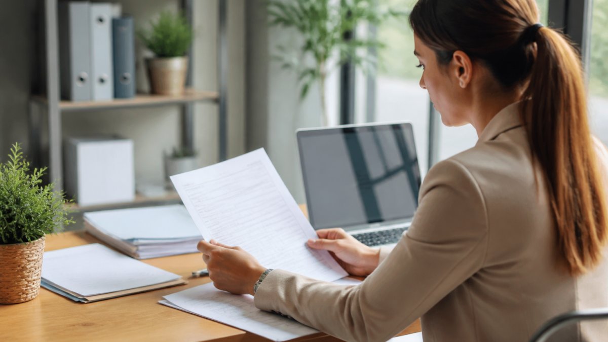 A property manager at a desk reviewing a tenant screening report on a laptop, with a rental application and pen nearby in a bright office